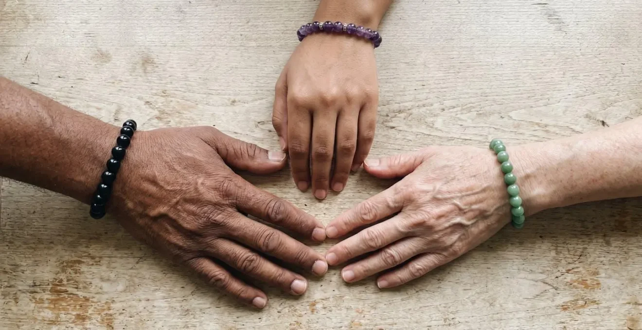Trois mains de couleurs de peau différentes portant des bracelets en pierres naturelles (obsidienne, améthyste, jade) sur un fond en bois