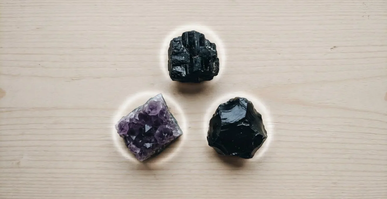 Trois pierres brutes (obsidienne, tourmaline noire, améthyste) disposées en triangle sur du bois clair avec un halo lumineux